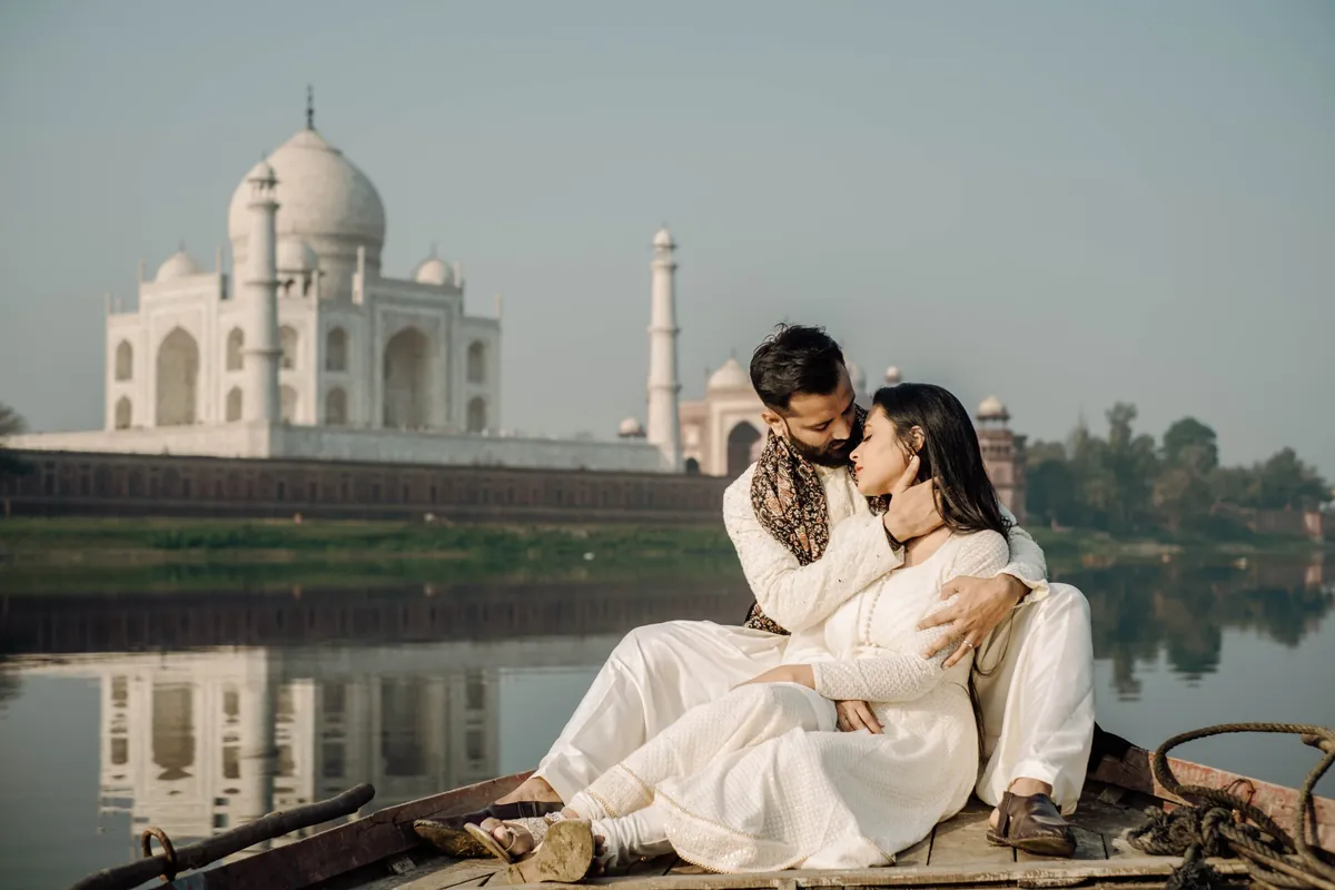 A couple in ivory sitting close on a wooden boat on the Yamuna, the Taj Mahal behind them
