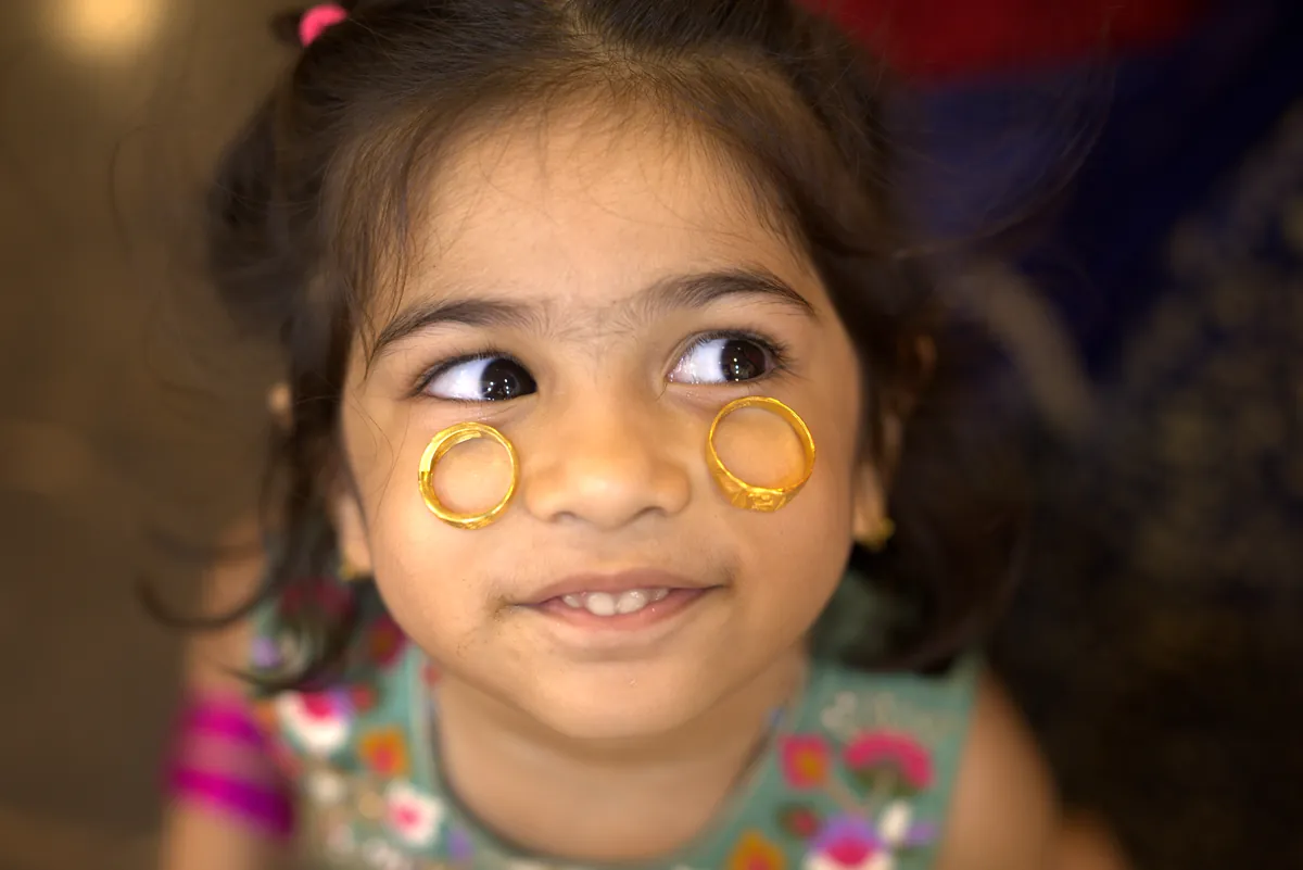 A little girl at a wedding with two gold rings balanced on her cheeks, eyes sliding sideways
